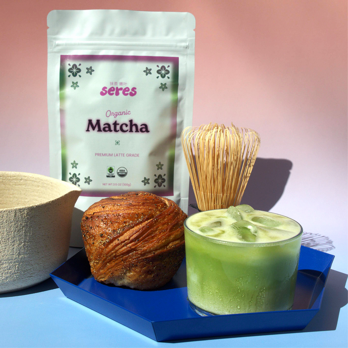 Seres Matcha Organic Daily 100g pouch next to an iced matcha latte, bamboo whisk, and fresh pastry on a tray.